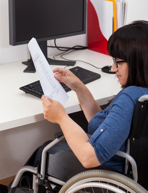 Vrouw in rolstoel met papieren in de hand zit aan haar bureau